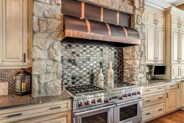 This kitchen has a rustic and natural feel, with stone finishes and warm wood accents. The cabinets are made of wood and have a distressed finish, which adds to the farmhouse-style aesthetic. The cabinet doors have a traditional panel design with visible knots and imperfections, contributing to the natural and rustic look of the kitchen. The countertops are made of natural stone, which adds a unique and organic element to the space. The backsplash is made of split octocagnol with a matte finish, which creates a subtle and neutral backdrop for the kitchen.