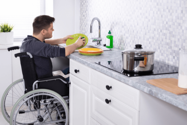 The image depicts a modern, fully accessible kitchen in a home setting. The kitchen features lower countertops, a sink, an oven, a refrigerator, and cabinets that are all within easy reach for individuals using wheelchairs. The space is well lit and designed with neutral tones, creating a welcoming and inclusive environment. The flooring appears to be smooth and flat, facilitating easy movement for mobility devices. This image demonstrates thoughtful design that emphasizes accessibility and inclusivity.