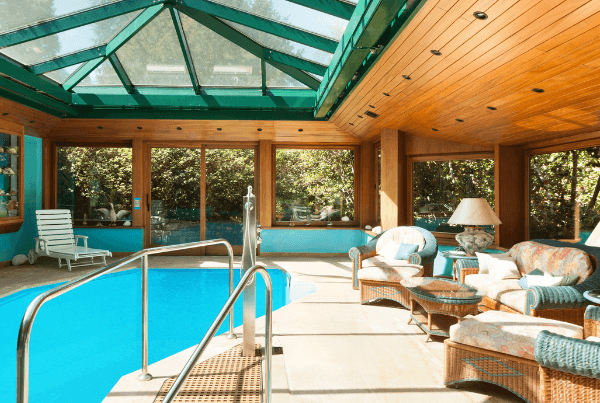 This image showcases an indoor swimming pool addition to a home. The pool is long and rectangular, filled with clear blue water. It's encased in a large, well-lit room with floor-to-ceiling windows along one side, allowing for ample natural light and offering a panoramic view of the outdoor landscape. The room's ceiling features exposed wooden beams, adding a rustic charm to the otherwise modern space. The pool deck is finished with light-colored tiles, which complement the pool's blue water. On one end of the pool, there's a set of built-in steps leading into the water, facilitating easy entry and exit. Adjacent to the pool, there's a comfortable seating area with two armchairs and a small table, providing a space for relaxation. A few potted plants are placed around the room, adding a touch of greenery and enhancing the connection to the outdoors. In summary, this image depicts a spacious and well-lit indoor pool addition, blending modern design with natural elements to create a serene and inviting environment for leisure and relaxation.