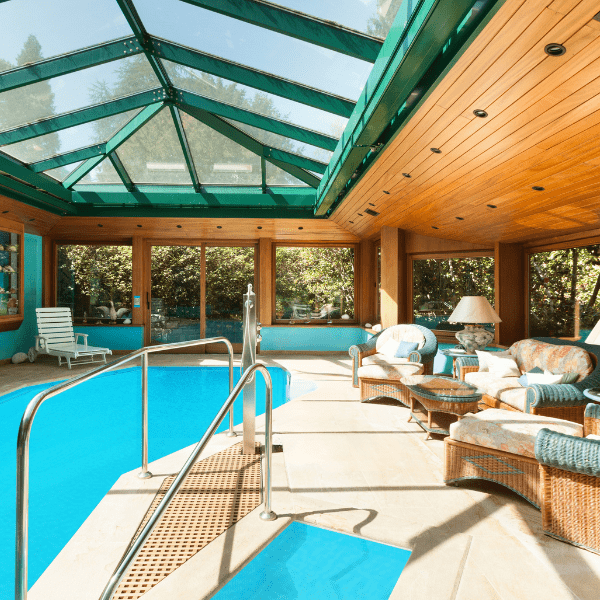 This image showcases an indoor swimming pool addition to a home. The pool is long and rectangular, filled with clear blue water. It's encased in a large, well-lit room with floor-to-ceiling windows along one side, allowing for ample natural light and offering a panoramic view of the outdoor landscape. The room's ceiling features exposed wooden beams, adding a rustic charm to the otherwise modern space. The pool deck is finished with light-colored tiles, which complement the pool's blue water. On one end of the pool, there's a set of built-in steps leading into the water, facilitating easy entry and exit. Adjacent to the pool, there's a comfortable seating area with two armchairs and a small table, providing a space for relaxation. A few potted plants are placed around the room, adding a touch of greenery and enhancing the connection to the outdoors. In summary, this image depicts a spacious and well-lit indoor pool addition, blending modern design with natural elements to create a serene and inviting environment for leisure and relaxation.