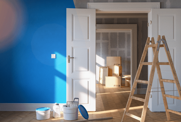 This image showcases an interior wall painting project in progress. The scene depicts a room with one wall partially painted in a bright, bold color. A paint roller attached to a long handle is seen mid-stroke, applying a swath of vibrant paint to the wall. A paint tray filled with the same color is visible on the floor below, with a drop cloth spread beneath it to protect the flooring from potential drips and spills. The room appears to be well-lit and features hardwood flooring. The other walls and ceiling in the room are painted in a light, neutral color, which provides a stark contrast to the wall being painted. In summary, the image captures a moment in the process of transforming a room through the application of bold, colorful paint to an interior wall.