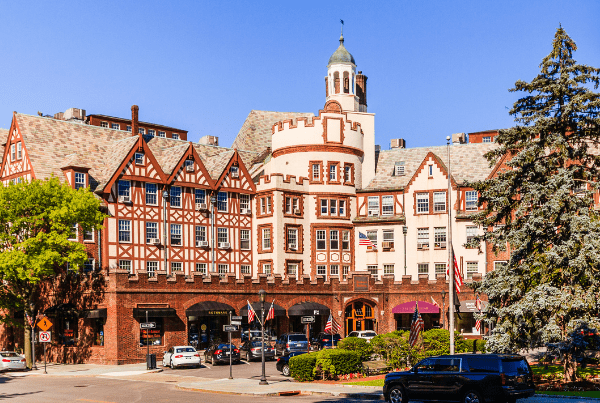This image depicts a serene residential neighborhood in Scarsdale, NY. The view presents a quiet, tree-lined street with well-maintained houses set back from the road. Each property appears to have a spacious front yard, enhancing the sense of privacy and space. The houses are mostly two-story, featuring a variety of architectural styles including colonial and Tudor. They are set against a backdrop of lush greenery, with mature trees dotting the landscape and providing shade. The street is clean, and there are sidewalks on both sides, making it pedestrian-friendly. The weather appears to be clear with a bright blue sky, suggesting a sunny day. Overall, the image presents a charming and peaceful suburban neighborhood, offering a high standard of living and a strong sense of community.