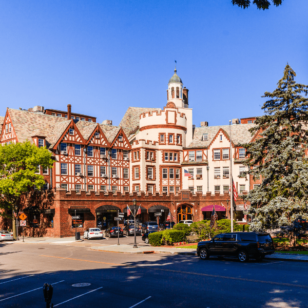 This image depicts a serene residential neighborhood in Scarsdale, NY. The view presents a quiet, tree-lined street with well-maintained houses set back from the road. Each property appears to have a spacious front yard, enhancing the sense of privacy and space. The houses are mostly two-story, featuring a variety of architectural styles including colonial and Tudor. They are set against a backdrop of lush greenery, with mature trees dotting the landscape and providing shade. The street is clean, and there are sidewalks on both sides, making it pedestrian-friendly. The weather appears to be clear with a bright blue sky, suggesting a sunny day. Overall, the image presents a charming and peaceful suburban neighborhood, offering a high standard of living and a strong sense of community.